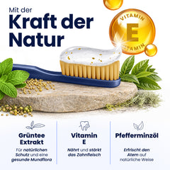Dental brush with toothpaste on a stone surface, surrounded by mint leaves and vitamin E symbol, with text in German about natural ingredients.